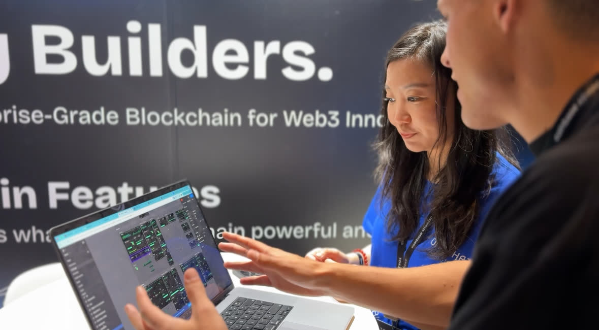 Two people leaning over a laptop at a conference stand, one of them gesturing at the screen.